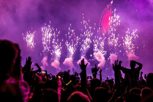 A lively crowd at a rave concert, with colorful fireworks lighting up the night sky in the background.